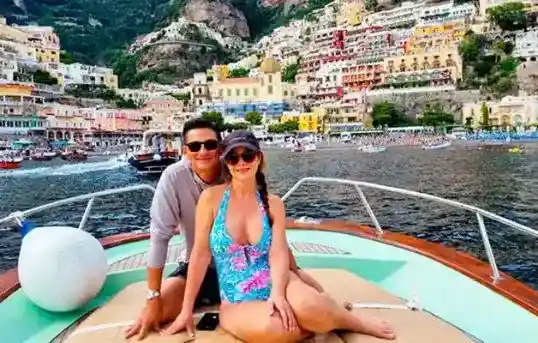A smiling couple relaxing on the spacious sundeck at the bow of the Gozzo Sorrentino boat Bellavita, with the cascading colorful houses of Positano in the Amalfi Coast visible on the cliffs behind them during a private tour with Frasil Charter.